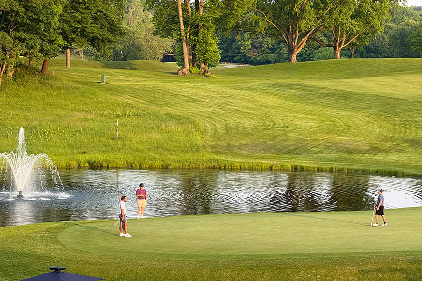 Image of a golf course with a pond and water fountain. Three golfers are also pictured.