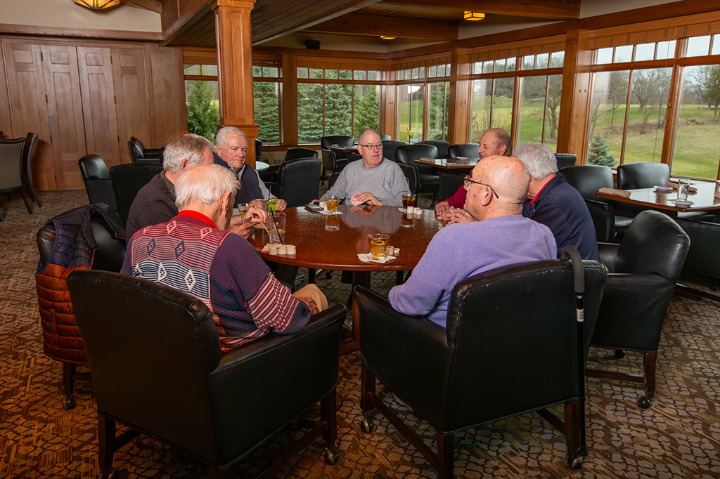 Members gathered around a table enjoying conversation and drinks inside the West Bend Country Club dining room with large windows overlooking the golf course.